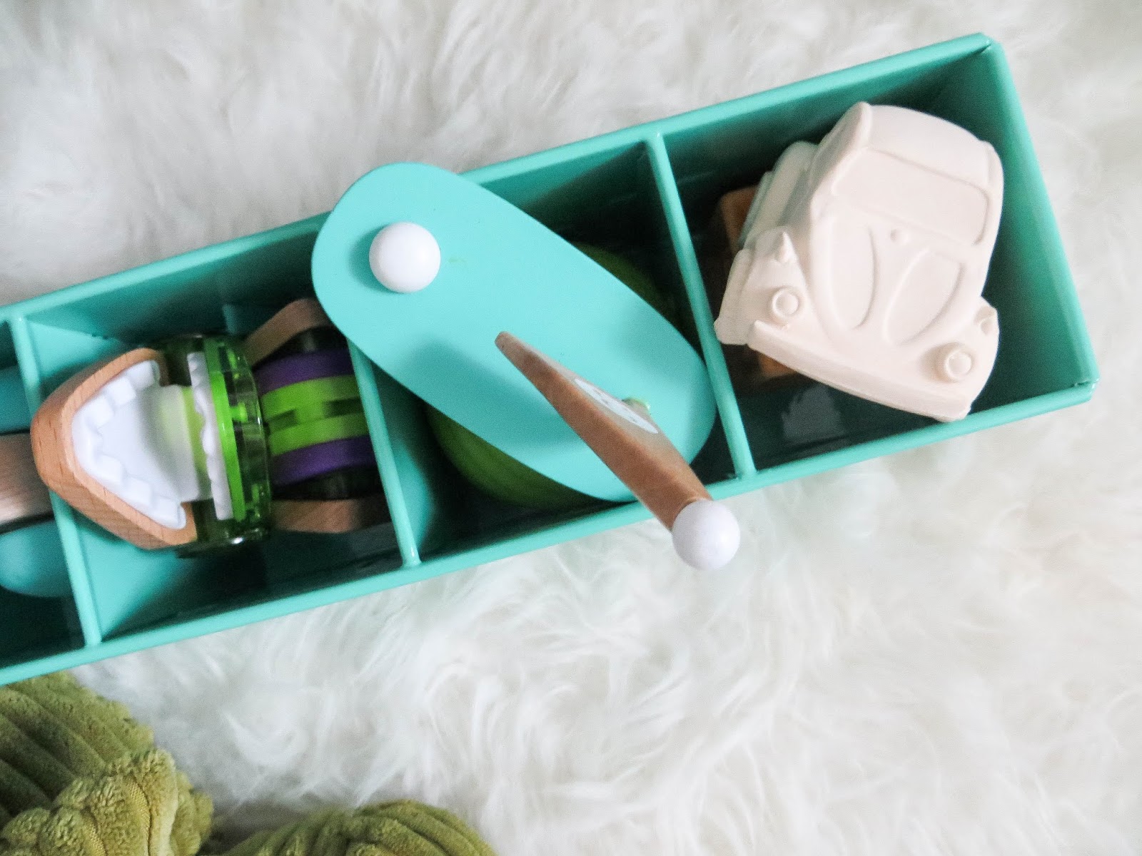 Overhead view of a teal divided tray holding small wooden and plastic toys on a white fluffy rug, soft natural light