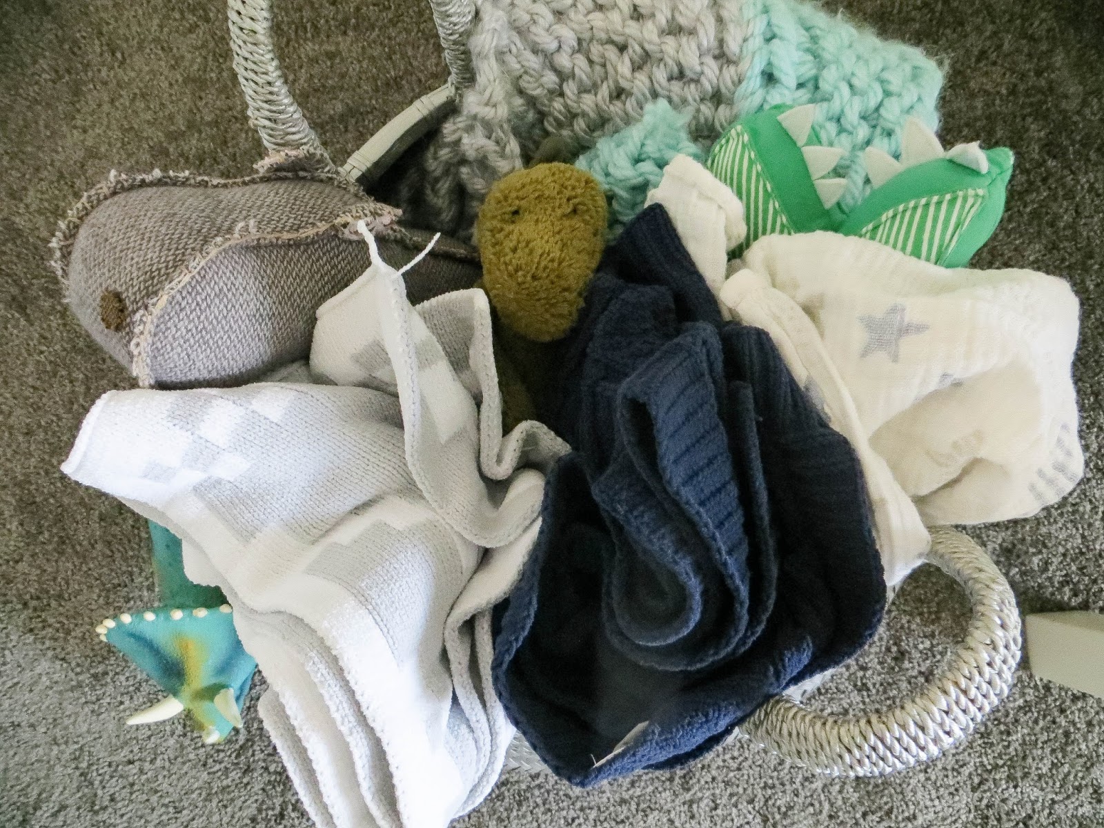 A woven basket filled with folded knit blankets, a plush toy, and soft baby items on a carpeted floor