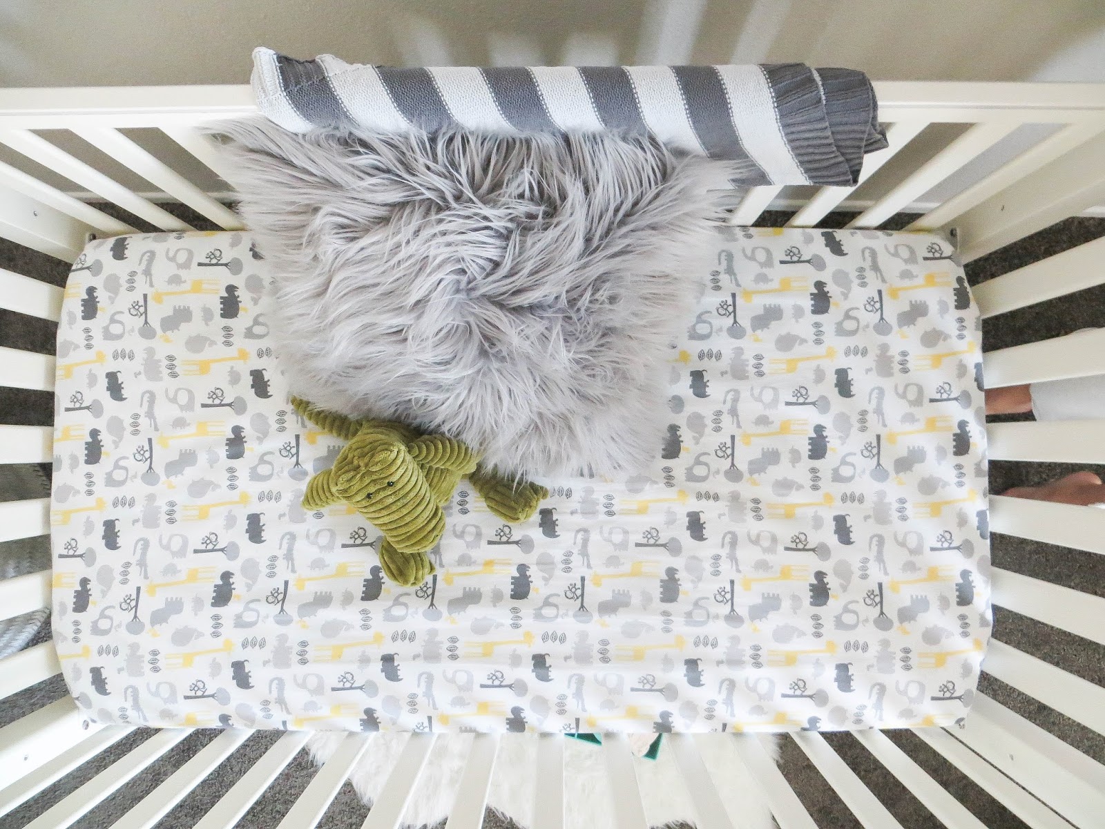 Overhead view of a white crib with an animal-print fitted sheet, a gray faux fur pillow, striped blanket, and green plush toy