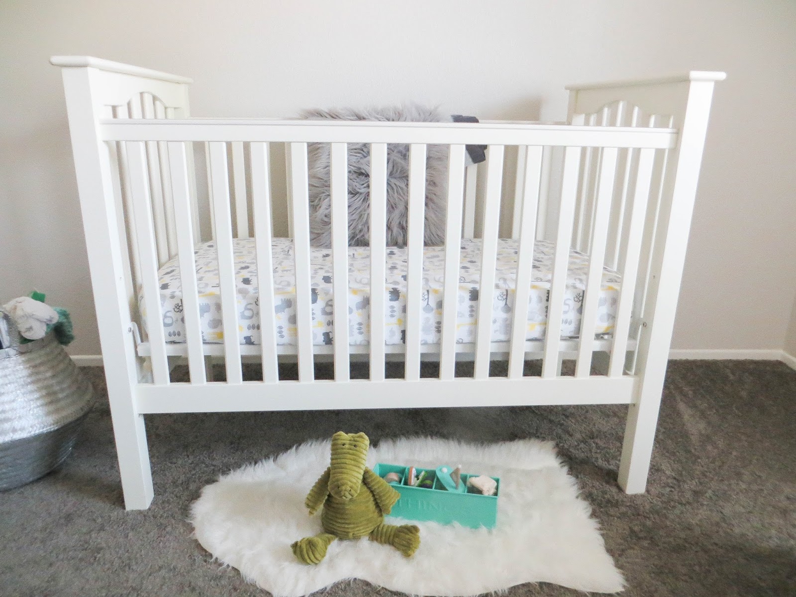 A white wooden crib with patterned sheet and gray fur pillow, with a green plush toy and teal storage bin on a faux fur rug
