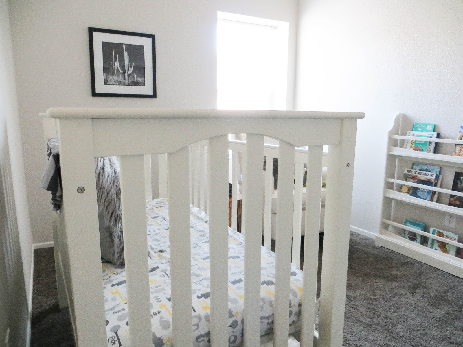 A white crib in a nursery with a patterned fitted sheet, framed black-and-white cactus art on the wall, and a bookshelf nearby, soft warm light filling the room