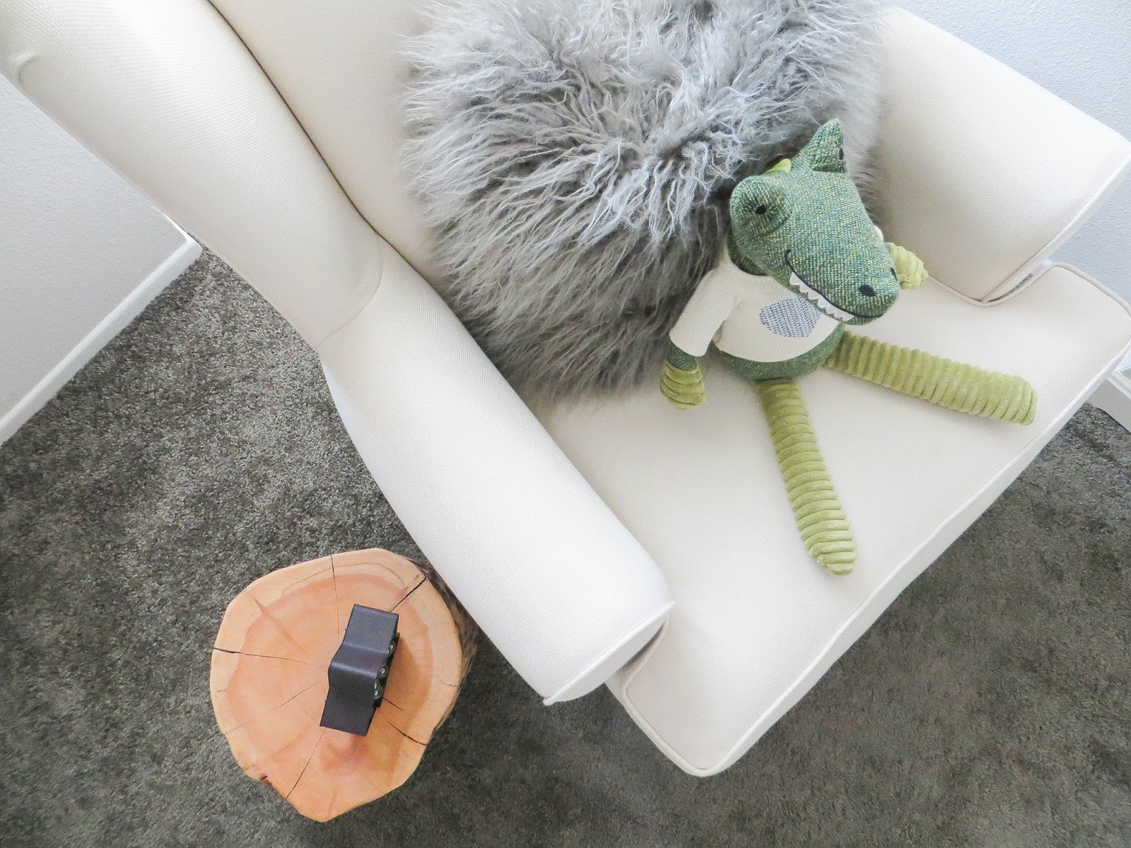 Overhead view of a white chair holding a round gray fuzzy pillow and a green stuffed toy animal, with a wooden stump side table, warm and cozy nursery vibe